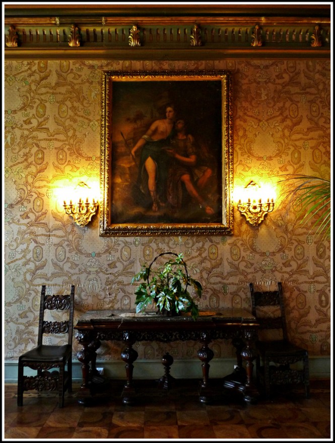 The heavy, dark wood with its intricate, ornate carvings,  is a prime example of castle furniture. The richly adorned wallpaper, wall candles, and medieval-era painting add to the castle theme.  Photo Credit