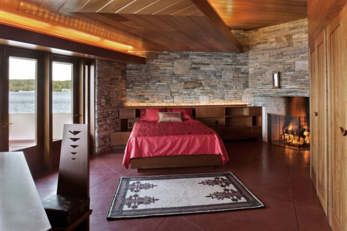 The master bedroom  features a rich wood, offset chevron ceiling design lit from above. It anchors itself into a rugged rock wall at the back of the room.