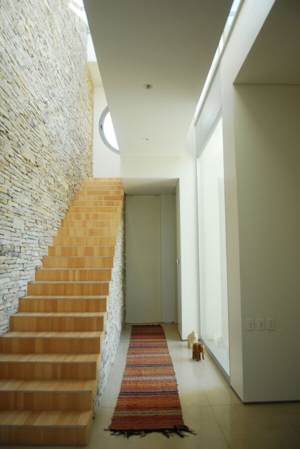 A wall of locally cut stone acts as a dramatic partition in the open plan living room and the primary material in the tactile twin bathroom. It continues into the organic, lightly wooded stairwell, which, without railings or glass panels, makes for a sleek design choice in a home where the safety of children, need not be considered. The stone is also a frequent reference for the warmly muted color palette used throughout Agua House’s interior.
Dominated by stone as they are, internal spaces incorporate neutral colors and blonde wood in an effort to retain and reflect as much natural light as possible. External spaces, with the reflection of the pool and an aspect that enjoys ample sun, prefer a chic combination of dark wood and chrome that contrasts with the subtropical landscape and subsequently lush gardens.