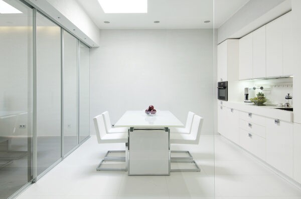 One could be forgiven for branding the heart of this space 'open plan,' but the versatile glass walls act, not only, to define the space, but also set the tone for the ambiance generated by the entire project. They speak to the high shine finish of the white kitchen cabinetry, the metallic light installation that crowns the neutrally soft living area and hovers directly over its glass coffee table, are reflected in the glass that frames the recessed, wooded fireplace and paid tribute to in the miniature greenhouse niche that graces the bathroom.