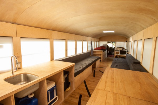 This photo shows the entire length of the converted bus home. The furniture is simple wood and the floor is made of reclaimed gym flooring. None of the structures built go above the window line keeping it linear and open to outside light.