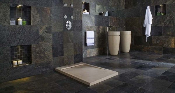 This incredibly large bathroom may be covered floor to ceiling in dark stone tiles, but it is grand and luxurious space. There is no separation for the shower and wall cubicles with recessed lighting provide shelving for bath products. The pedestal sinks brighten up the color palette.